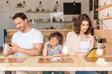 parents using gadgets while their son bored in cafe