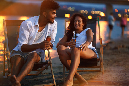 Young mixed race couple sitting and relaxing  at the beach on beautiful evening.They sitting on the sunbeds and drinking bear.