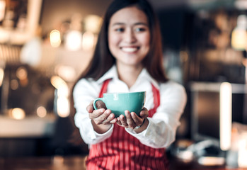 Close up on coffee up with blur asian woman barista holding mug with two hand with smiling face at cafe bar background,Focus on coffee up