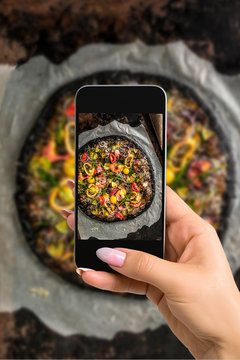 Photographing food concept - woman takes picture of italian pizza with black dough and seafood on a baking tray from the oven