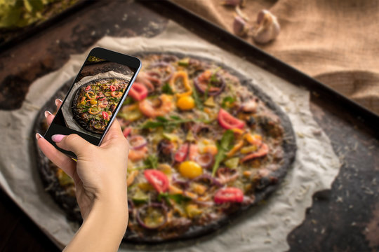 Photographing food concept - woman takes picture of italian pizza with black dough and seafood on a baking tray from the oven