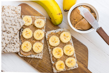 Healthy vegan snack with scandinavian rye crispbread, homemade peanut butter and slices of Canary island bananas bananas, wood cutting board, top view