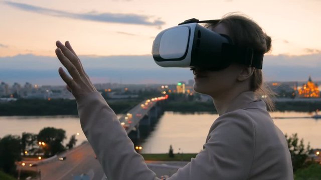 Woman uses virtual reality glasses in the city after sunset. Evening time, twilight. Relax, entertainment and technology concept