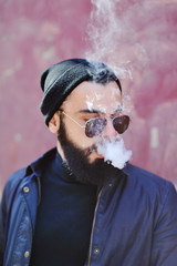 A young bearded man in a black hat and sunglasses smokes an electronic cigarette or vape