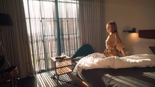 Young caucasian woman in brown beige robe streches out hands sitting on bed in the morning then walks to look through big window. Shot with Sony a7s and Atomos Ninja Flame.