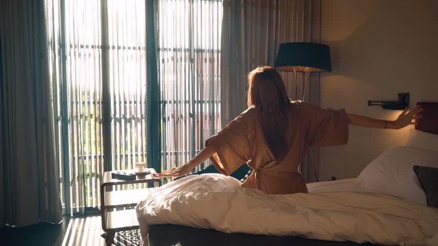 Young caucasian woman in brown beige robe stretches out hands sitting on messy bed in the morning in front of big window. Shot with Sony a7s and Atomos Ninja Flame.