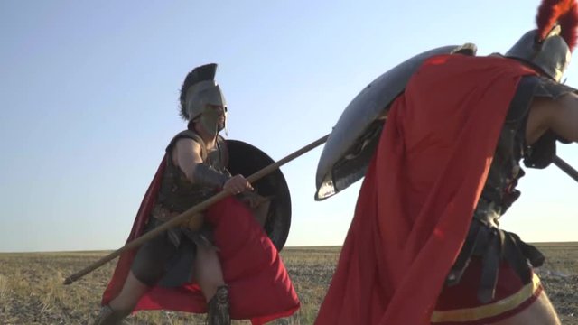 Roman gladiators in heavy armor and red cloaks beat on spears and shields in the field, slow motion shooting
