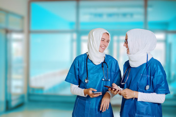 Two female middle eastern doctors in blue medical uniform standing in a hospital hallway, looking at each other, laughing while holding smart phones in their hands