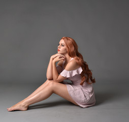 Naklejka premium full length portrait of a pretty girl with long copper hair, wearing a simple purple dress. seated pose against a grey background.