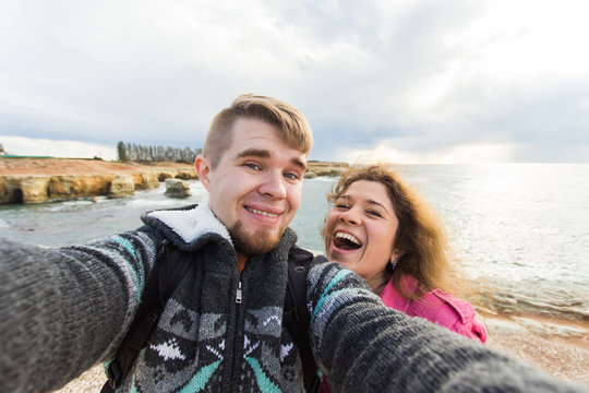 Happy traveling couple in love taking a selfie on phone at the beach on winter or autumn day. Pretty girl and her handsome boyfriend having fun, crazy emotional faces , laughing.
