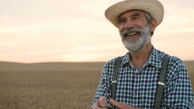 Close up portrait of old happy fermer with beard in the hat smiling in the golden field on the colorful sky background.