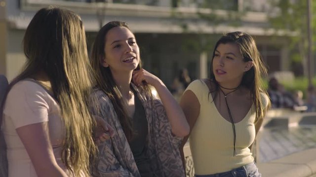 Group Of Multiethnic Friends Lean Against Fountain In City Square, Girl Brushes Her Friend's Hair Out Of Her Face, They Laugh