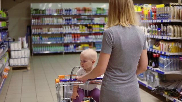 Young mother with her little baby sitting in a grocery cart in a supermarket is walking forward. The kid is looking around and waving her hand