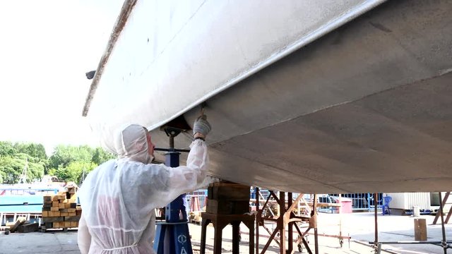 Worker paints metal bottom of ship at shipyard in port of Moscow. Process of repair and reconstruction of sea vessel. Outdoor work. Technology of manual painting boats. Industry of water transport.