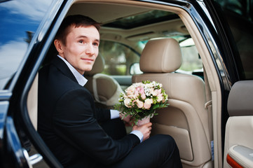 Portrait of a handsome groom sitting in the wedding car with a bouquet in his hands.