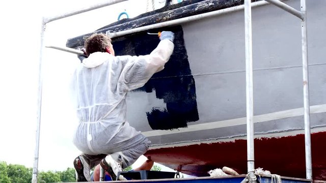 Worker paints metal of old rusty ship at shipyard in port of Moscow. Process of repair and reconstruction of sea vessel. Outdoor work. Technology of manual painting boats. Industry of water transport.