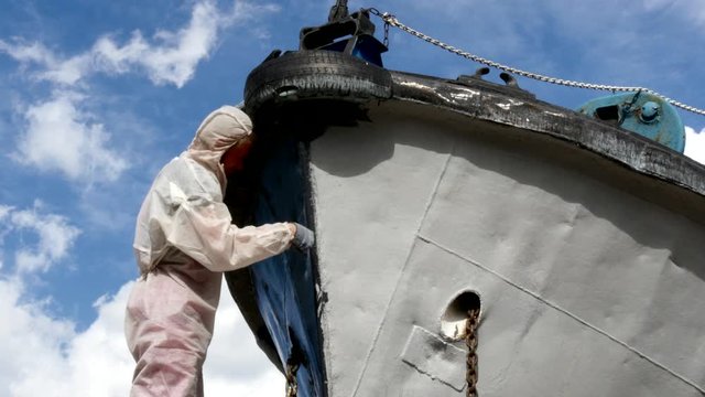Worker paints with brush metal front of ship at shipyard in port of Moscow. Process of repair of sea vessel. Outdoor work. Technology of manual painting boats. Industry of water transport.
