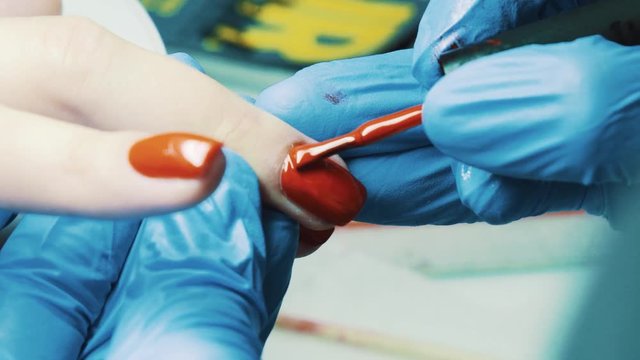 Beautician hand in blue rubber gloves paints red nail polish using brush on client thumb nail in bright salon, close up