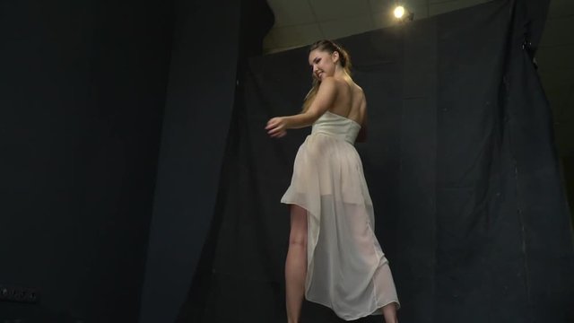 Cheerful girl in white dress turns on camera and smiles, shooting in studio. Slow motion