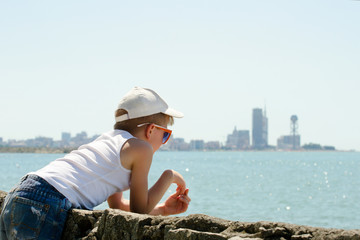 Boy in a cap looks at the sea. Side view. City in the distance.