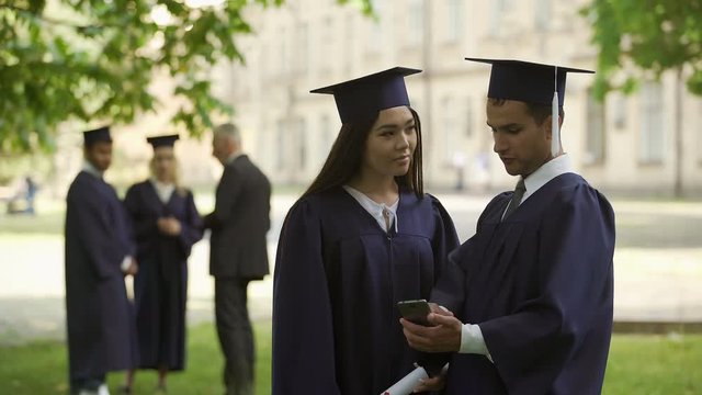Male graduate showing something on mobile phone to female classmate, application