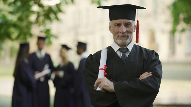 Senior man in academic regalia holding diploma, education at any age, new degree