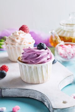 Colorful cupcakes with buttercream and fresh berries on cutting board. Tea pot with green tea and pink rose sugar on background. Closeup view, toned image