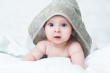 Cute baby girl or boy after shower with towel on head in white sunny bedroom. Child with big blue eyes relaxing in bed after bath