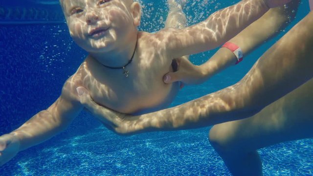 11 months old baby boy learns to dive. Mom's hands support him. Surprised looking with open eyes underwater. Slow motion 10 bit video