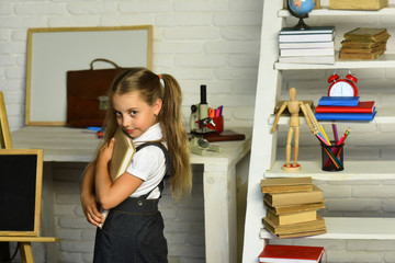 Schoolgirl with ponytails holds book on light classroom background