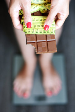 Young woman on weighting scale holding chocolate bar and measurement tape