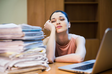 A young woman sits in front of a pile of papers and a computer holding her head