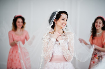 Fantastic bride posing in wedding dress with helpful pretty bridesmaids in big white room.