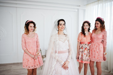Fantastic bride posing in wedding dress with helpful pretty bridesmaids in big white room.