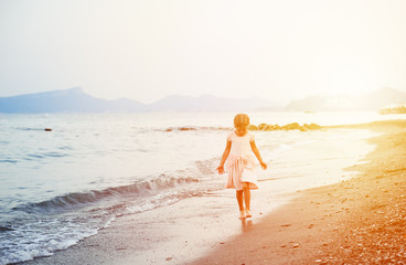 Little girl in summer dress traveling alone on coastline at sunset. 