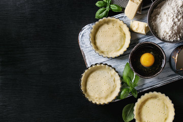 Dough for baking quiche tart in small baking form ready for bake. Ingredients above flour, egg, cheese, basil, cream on white serving board over black burned wooden background. Flat lay, copy space