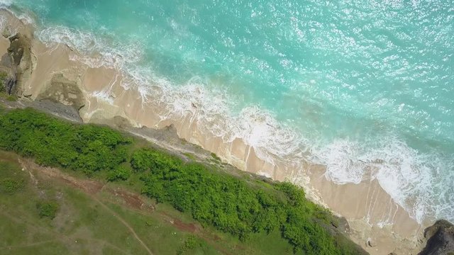 AERIAL TOP DOWN: Curly foamy whitewater ocean waves splashing gently against cliffy shore washing white sandy beach in sunny Bali island. Steep rocky crag wall rising above the crystal clear blue sea