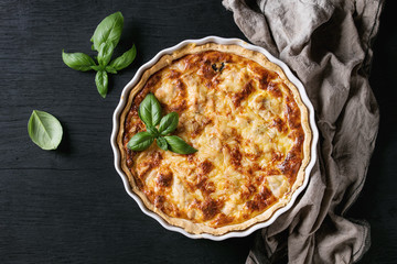 Baked homemade quiche pie in white ceramic form served with fresh greens and textile towel on black...