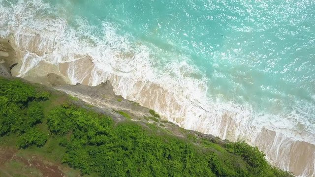 AERIAL TOP DOWN, CLOSE UP: Foamy whitewater ocean waves splashing gently against cliffy shore washing white sandy beach in sunny Bali island. Rocky crag wall rising above the crystal clear emerald sea