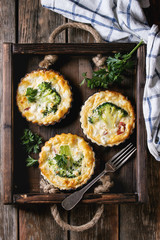 Baked homemade quiche pie in mini metal forms served with fresh greens, kitchen towel and fork in dark wood tray on old plank wooden background. Flat lay with space
