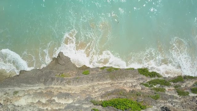 AERIAL TOP DOWN CLOSE UP: Curly foamy whitewater sea waves splashing gently against cliffy rocky shore washing white sand from the beach into the crystal clear ocean on sunny summer day in Bali island
