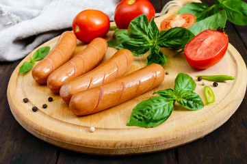 Juicy sausages with tomatoes and basil on a round cutting board on a dark wooden background.