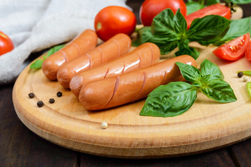 Juicy sausages with tomatoes and basil on a round cutting board on a dark wooden background.