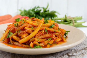 Spicy warm salad of green beans in tomato sauce on a plate on a white wooden background. Vegan dish.