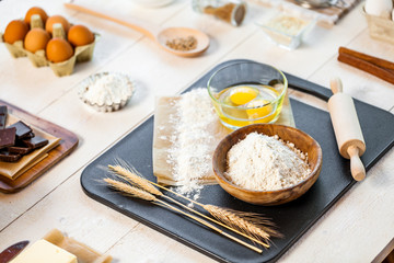 Baking ingredients in rural kitchen - dough recipe ingredients (eggs, flour, milk, butter, sugar) and rolling pin on wooden white table.