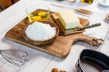 Baking ingredients in rural kitchen - dough recipe ingredients (eggs, flour, milk, butter, sugar) and rolling pin on wooden white table.