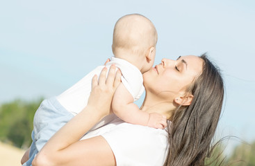 A small newborn baby boy and young pretty mother embracing him with fondness in wheat field at sunny summer day. Blue sky background