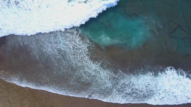 AERIAL TOP DOWN SLOW MOTION Foamy waves in crystal clear ocean breaking on coral reef on exotic beach of stunning Bali island. Turquoise sea splashing over rocky shoal in shallow water at the seashore