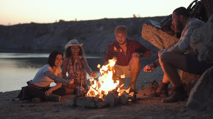Group of multiracial people sitting around campfire grilling marshmallows and having fun on coast.