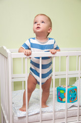 a small child looks up, standing in his baby bed
